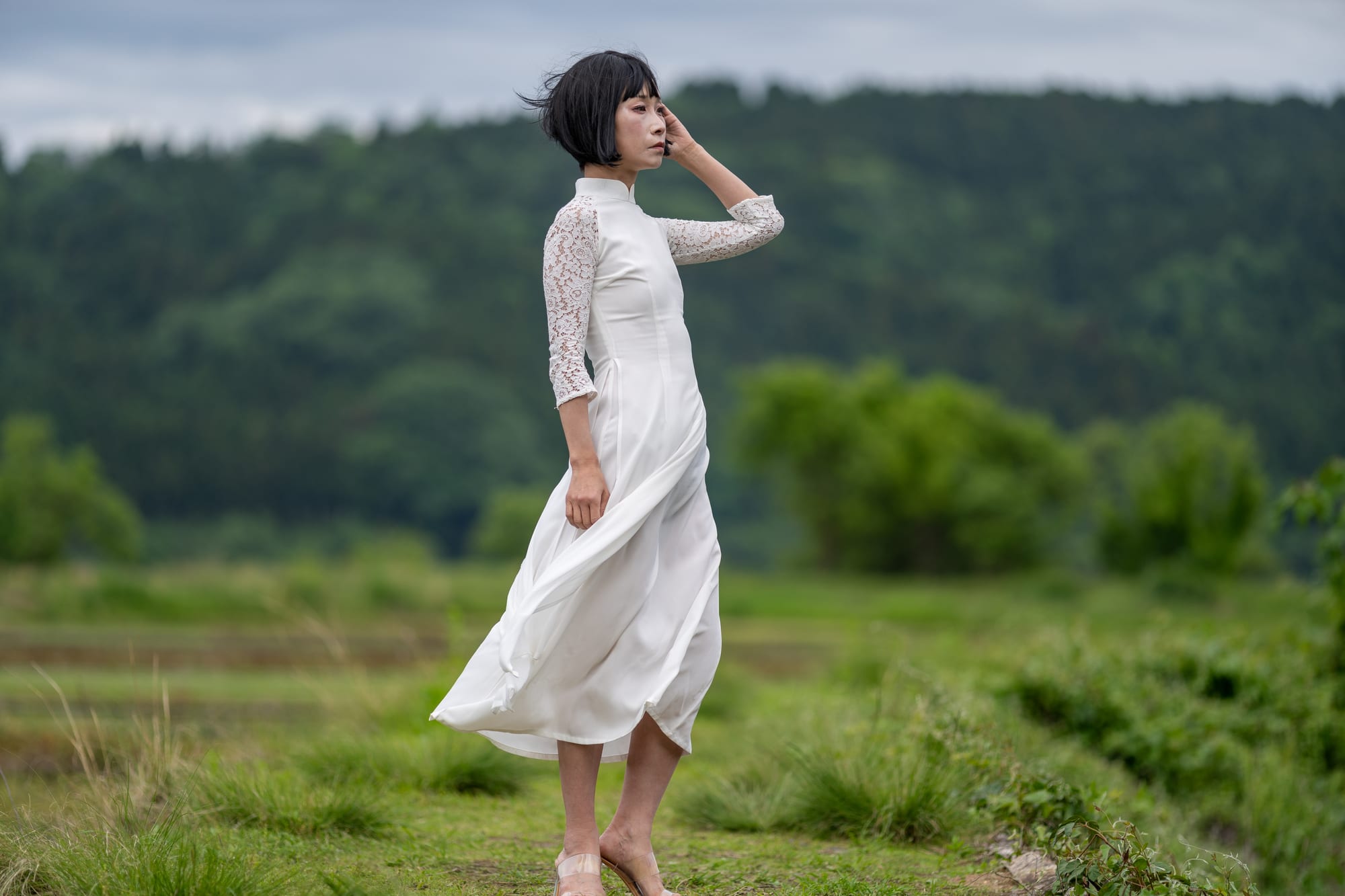 Tideline 05 — 田園を背に立つ白衣装の横顔。風に裾と髪が流れる。